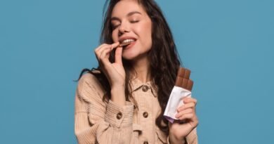 Foto colorida de mulher comendo um tablete de uma barra de chocolate. Ela segura uma barra de chocolate em outra mão - Metrópoles