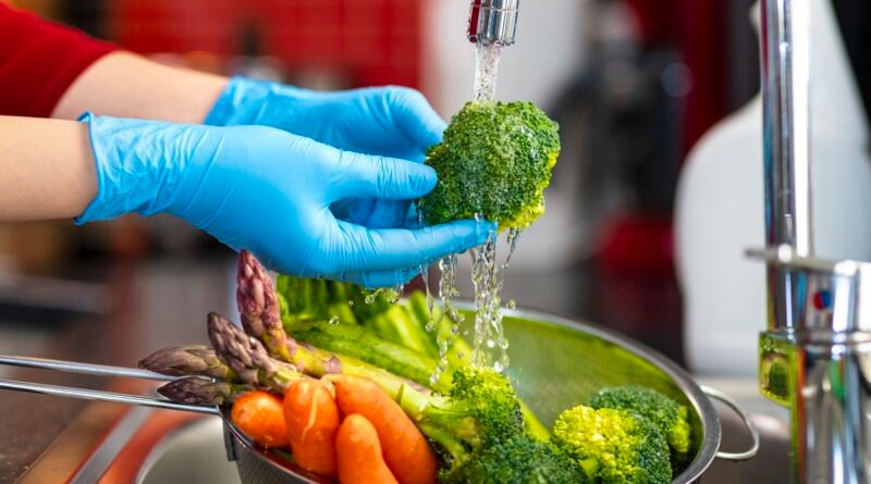 Imagem mostra as mãos de uma mulher usando luvas lavando alimentos em baixo de uma torneira - Metrópoles